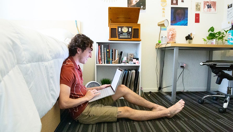 Student using laptop while sitting on the floor besides the bed in the dorms