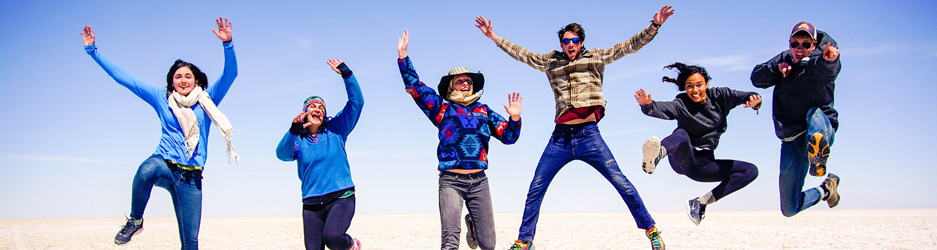 Six students and staff jumping in the air with the sand and sky in the background