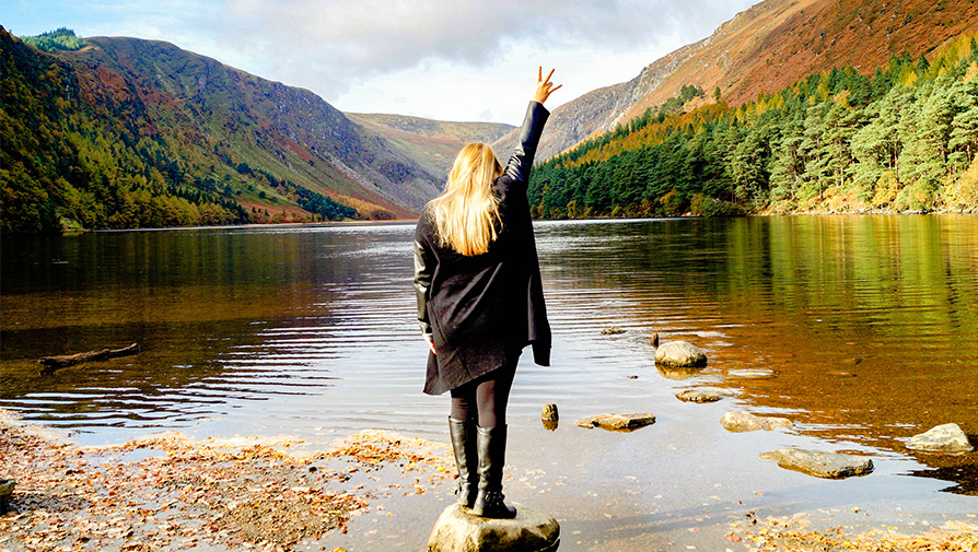 Female student posing the ASU pitchfork sign while looking down a beautiful river with hills on both sides