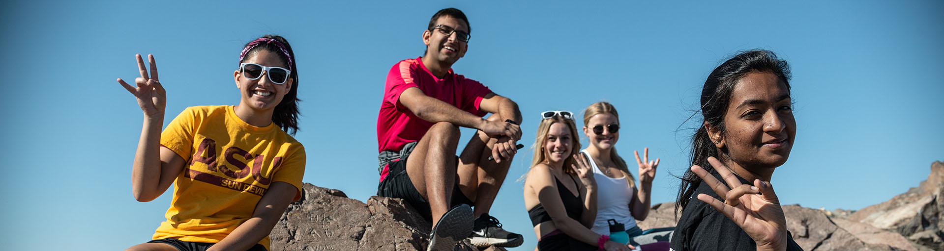 Five students posing with the ASU pitchfork while sitting on a large rock